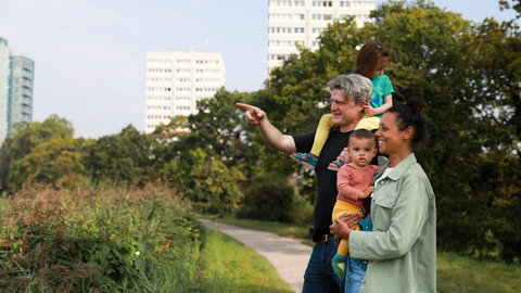 A family of four are at a nature reserve with buildings visible in the background. The father is pointing to show his partner and children something in the distance