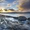 A coastal landscape, with the sea gently lapping at smooth rocks as the sun sets behind scattered clouds