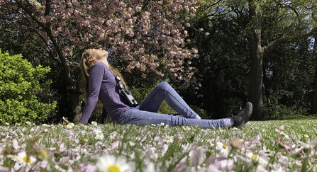 A beautiful young woman sits on the grass in a park, with daisies in the foreground and blossoming trees behind. She has her head thrown back and her eyes closed, enjoying the peace of nature