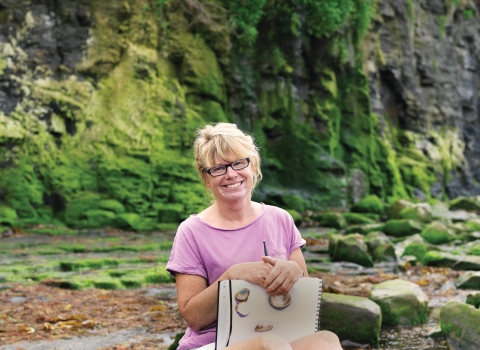 Carol sits on the beach with her sketchbook