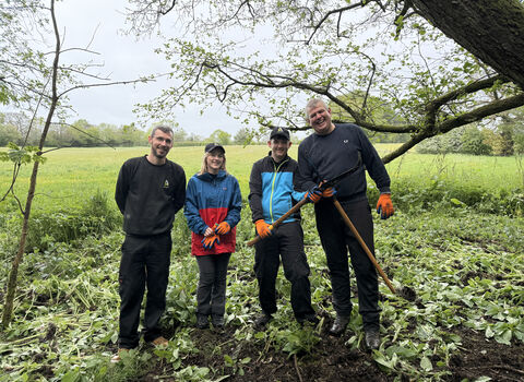 4 poeple standing beneath trees with tools and a field in the background